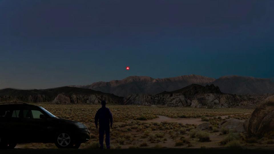 A digital rendering of a person standing in a desert landscape at night, with several red light sources visible in the sky.